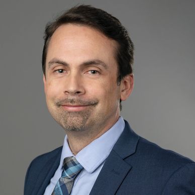 Professional man in a blue suit and patterned tie smiling with a neutral gray background.
