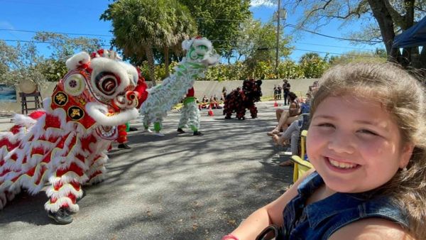 A smiling girl watches a colorful lion dance outdoors.