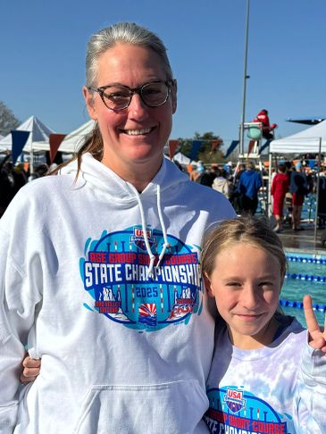 Two smiling females at a swim meet, wearing championship hoodies.