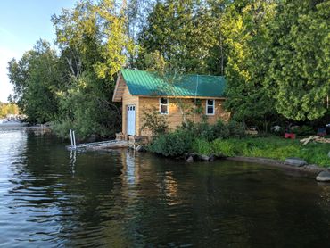 Wooden boathouse with green roof by the lakeside surrounded by trees.