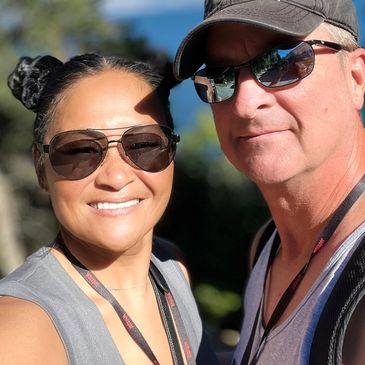 Smiling couple wearing sunglasses and lanyards, enjoying a sunny day outdoors.