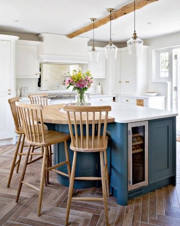 Bright kitchen with blue island, wooden chairs, and pendant lights.