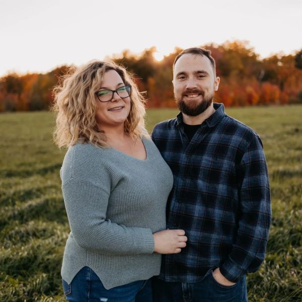 Smiling couple standing in a field with autumn trees in the background.