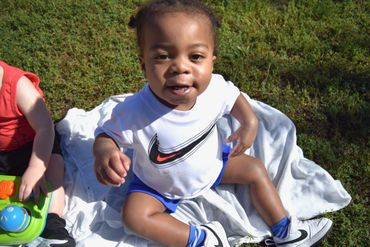 Smiling toddler sitting on a blanket outdoors, wearing a Nike shirt and white sneakers.