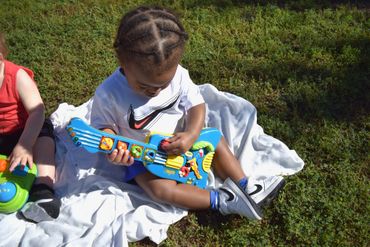Toddler playing with a colorful toy guitar on a blanket outdoors.