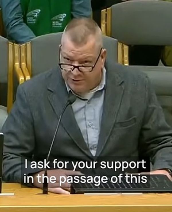 Man in glasses speaks at a formal meeting, asking for support.