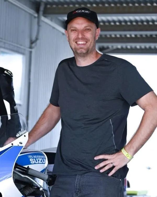 Smiling man in a black shirt and cap standing next to a blue Suzuki bike.