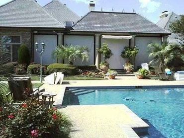 A backyard swimming pool with plants and a house in the background.