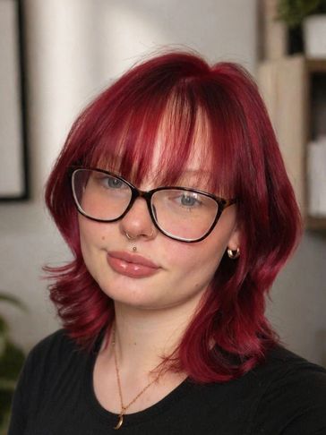 Young woman with vibrant red hair, glasses, and a septum piercing.