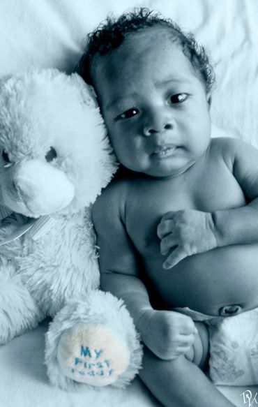 Newborn baby lying next to a teddy bear with "My First Teddy" on its paw.