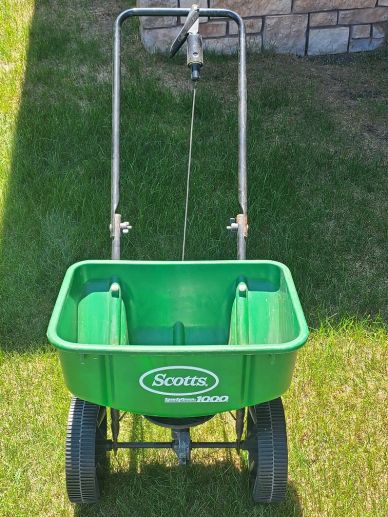 Green Scotts lawn spreader on grass with stone wall background.