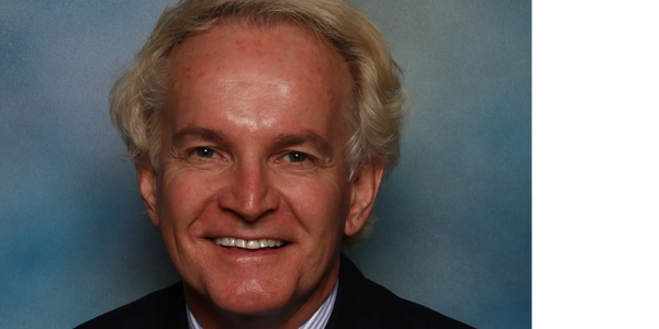 Smiling middle-aged man in a suit and tie against a blue-gray backdrop.