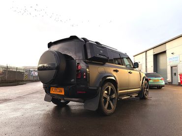 Matte black Land Rover Defender with urban accessories parked in an industrial area at sunset.