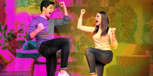A man and woman joyfully celebrate together indoors with raised fists and lifted legs.