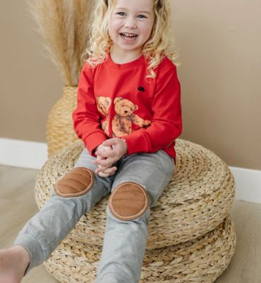 Smiling child in red sweatshirt and gray pants with knee patches sitting on a woven stool.