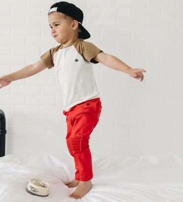 Young boy in red pants and backward cap jumping on bed.