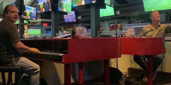 Two men playing red pianos in a lively venue with multiple TV screens.