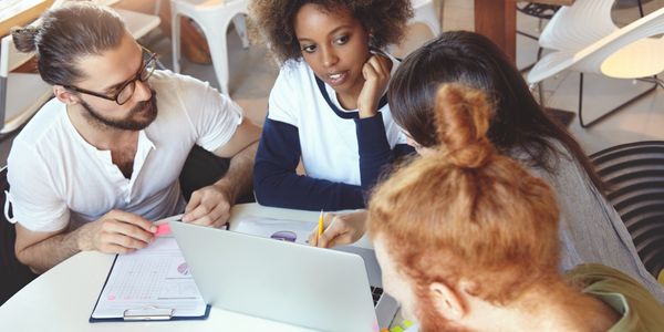 A diverse group collaborating on a project around a laptop in a casual workspace.