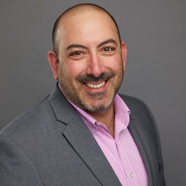 Smiling man in a gray blazer and pink checkered shirt against a gray background.