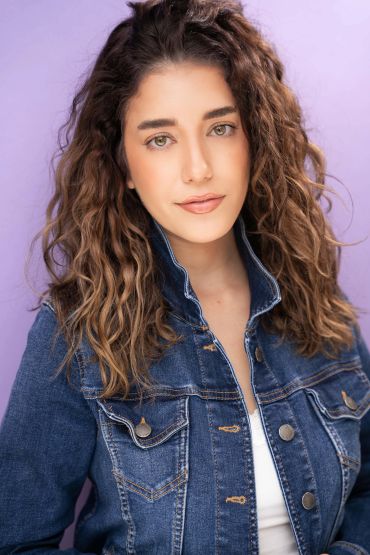 Young woman with curly hair in denim jacket against purple background.