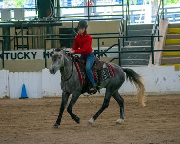 Young rider in red shirt rides a gray horse indoors.