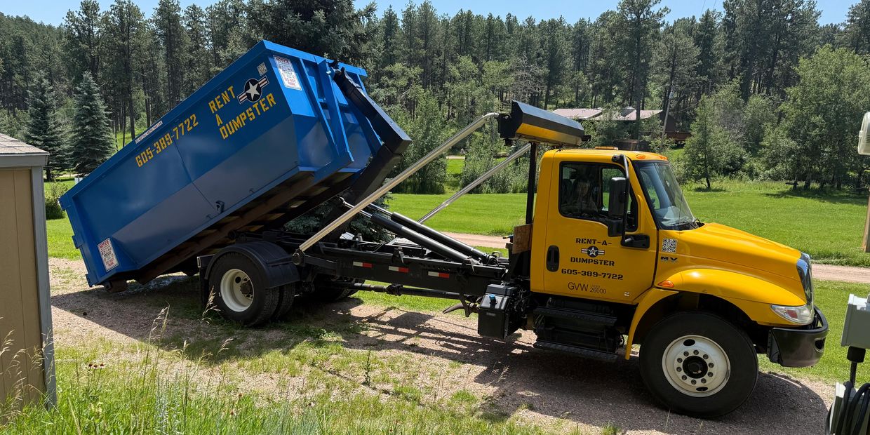 Yellow truck unloading a blue dumpster in a green, wooded area.