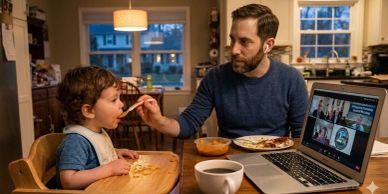 A father feeds his toddler at the dining table while attending a virtual meeting on his laptop.