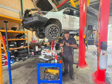 Mechanic working on car parts in a garage with a lifted white vehicle.