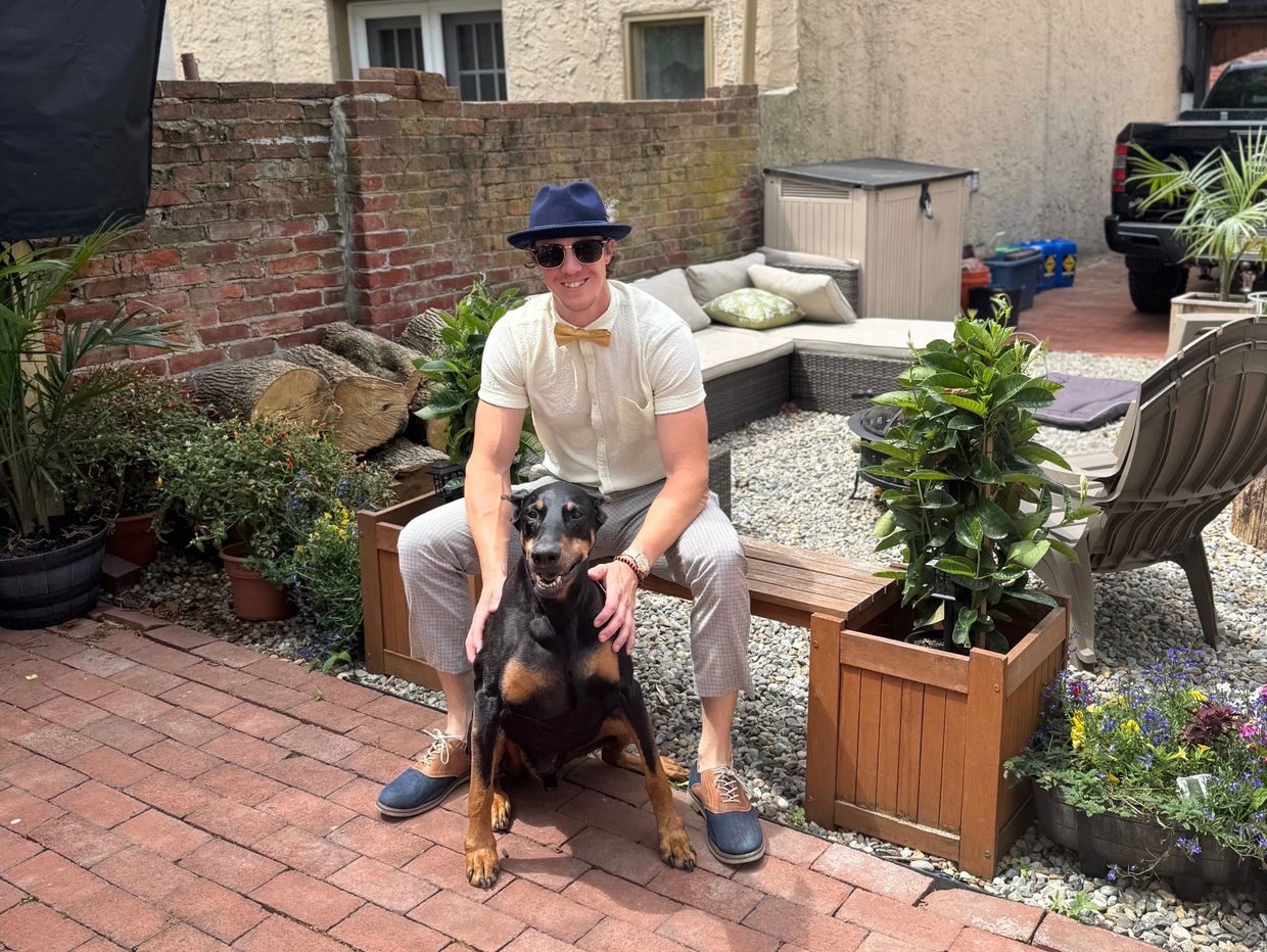 Man wearing a hat and sunglasses with a Doberman in a backyard.