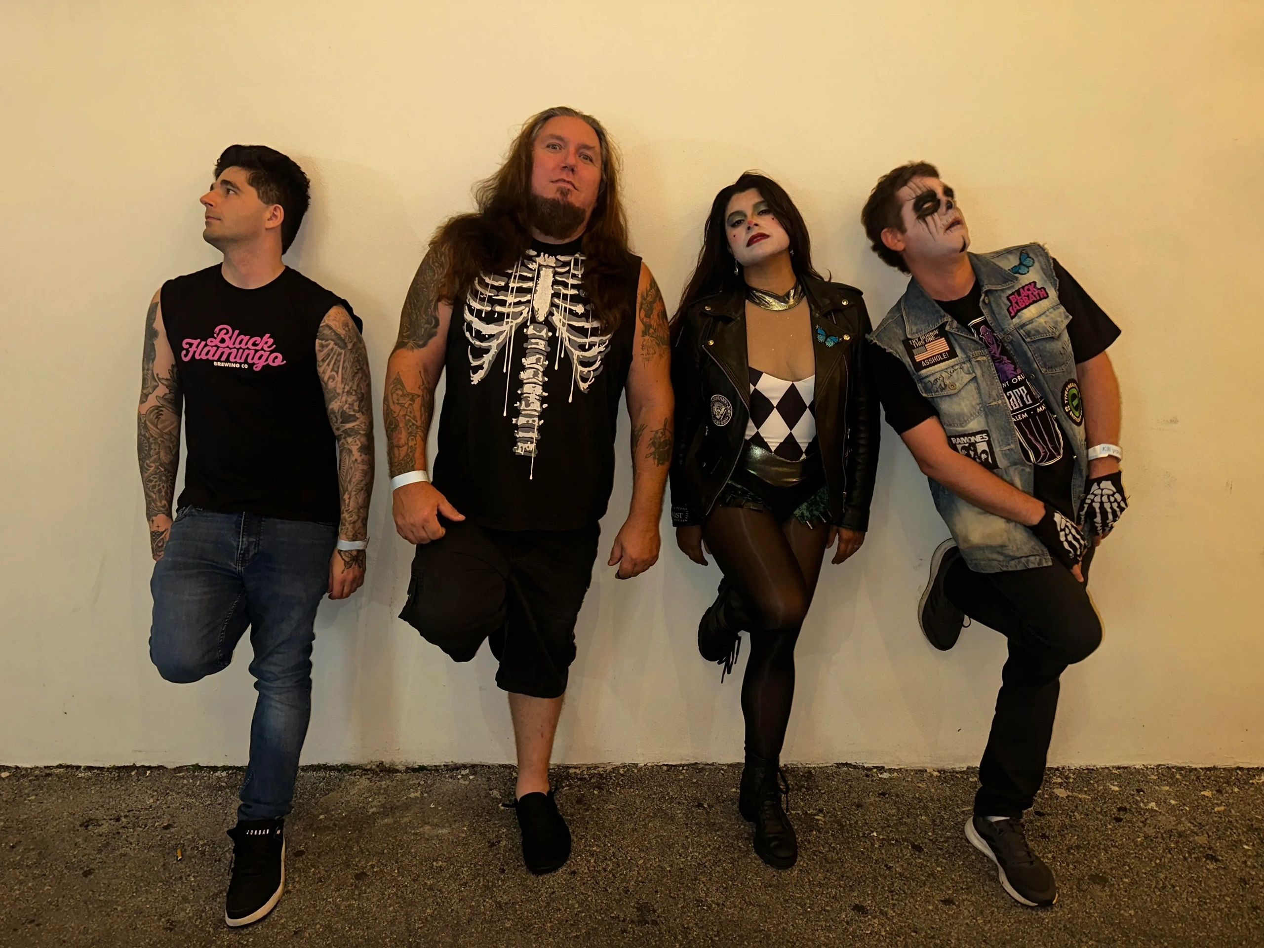 Four people posing against a wall, dressed in rock and gothic-inspired outfits with tattoos and face paint.