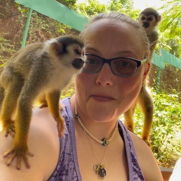 Woman with glasses and two small monkeys on her shoulders in a lush outdoor setting.