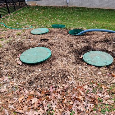 Green septic tank lids in a backyard with mulch and a blue hose.