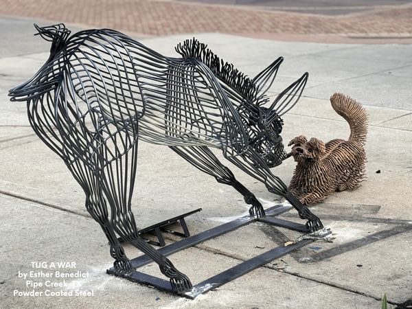 Wire sculptures of a cat and dog play tug of war on a sidewalk.