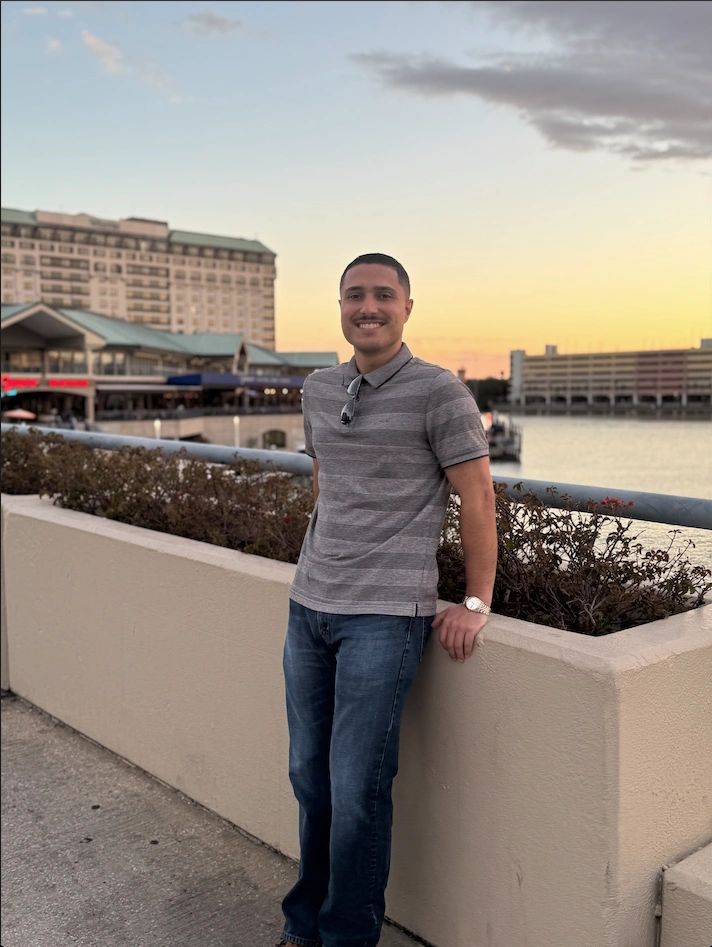 Man leaning on a concrete planter with waterfront and buildings behind.