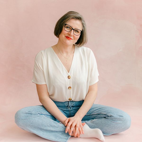 Woman with glasses sitting cross-legged on pink backdrop, smiling.