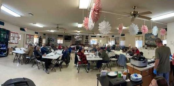 A community room with people seated at tables, decorated with hanging snowflakes and paper pom-poms.