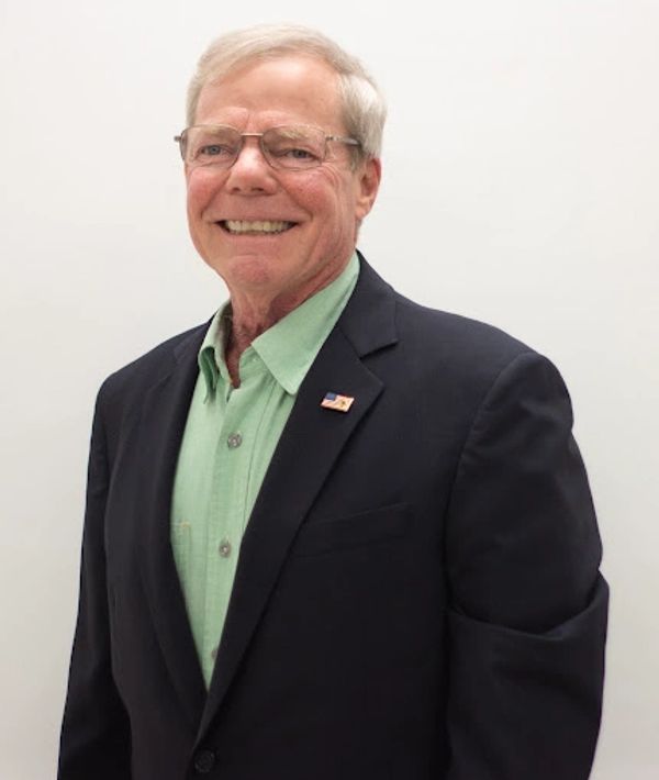 Smiling elderly man in glasses, green shirt, and dark blazer with an American flag pin.