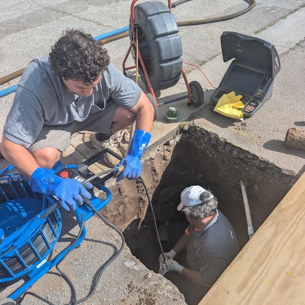 Two workers performing underground pipe inspection using specialized equipment.