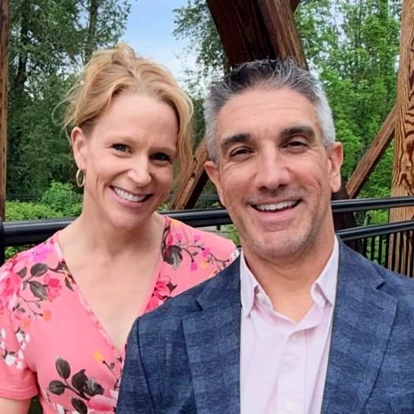 Smiling couple posing outdoors with greenery and wooden structure behind them.