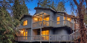 Modern cabin-style house with warm interior lights in a forested area at dusk.
