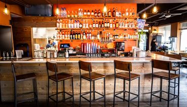 Modern bar with wooden stools and a vibrant orange wall filled with liquor bottles.