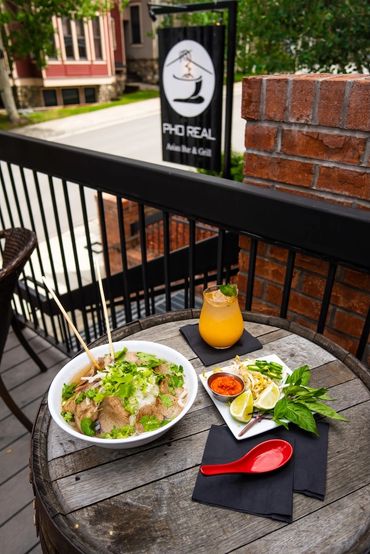 A bowl of pho with fresh herbs and a drink on a wooden barrel table outside a Vietnamese restaurant.