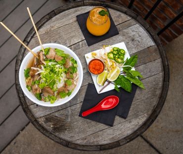 Bowl of pho with fresh herbs, lime, chili, and a refreshing orange drink on a rustic wooden table.