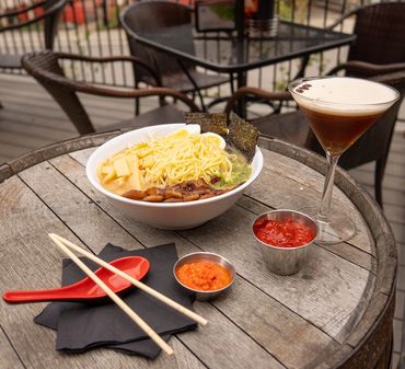 A bowl of ramen with chopsticks, sauces, and a cocktail on a rustic wooden table.