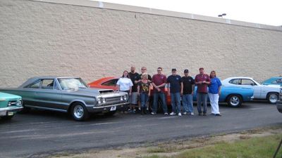 Group of people posing with classic cars in a parking lot.