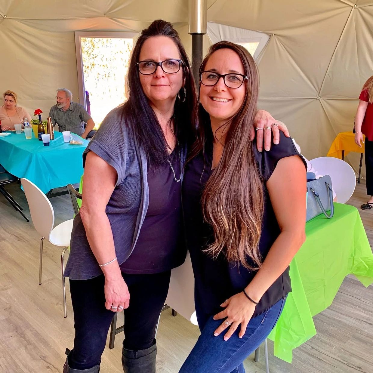 Two smiling women with glasses posing together indoors at a casual gathering.