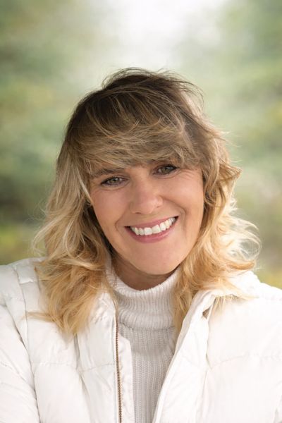 Smiling woman with blonde hair in a white jacket and sweater outdoors.