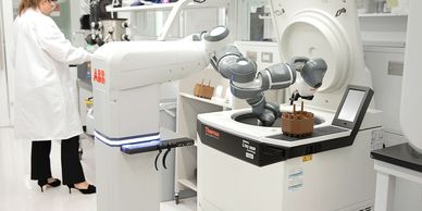 A robotic arm operates a laboratory centrifuge while a scientist works in the background.