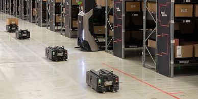 Robotic warehouse automation with small robots navigating aisles of shelves filled with boxes.