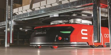 A red Swisslog warehouse robot navigating under metal shelves.